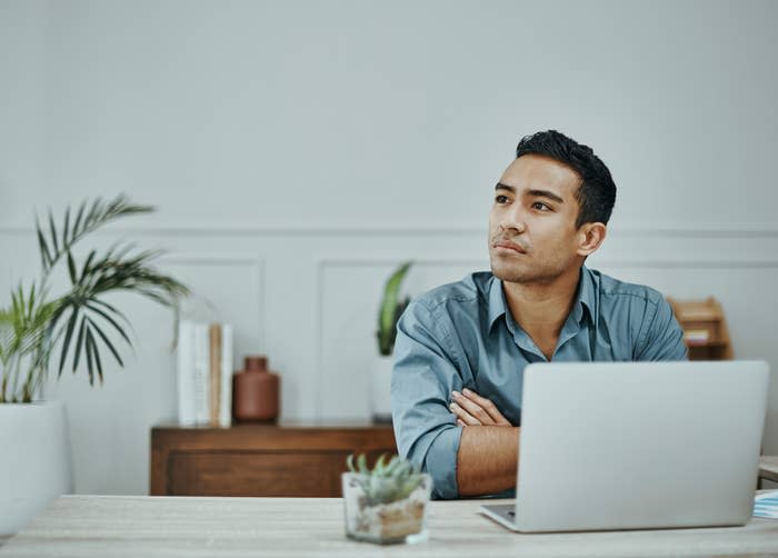 A man sits at a desk with a laptop, looking thoughtfully to the side. The background includes plants and a bookshelf