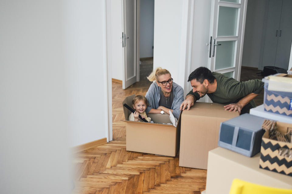 Family of three is moving in or out of a room in an apartment or a house. Mother and father are playing with the child on the floor of the room. The child is sitting in a box and holding what seems to be a vacuum cleaner stick. They are all smiling and the father is leaning towards the adorably cute kid.