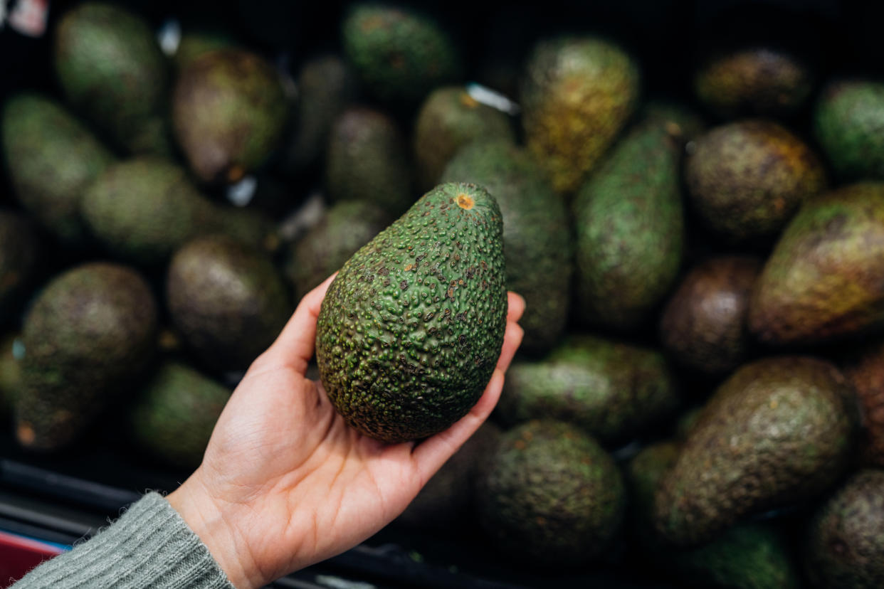 Frauenhand mit Avocado vor einem Supermarktregal voller Avocados. (Bild: Getty Images)