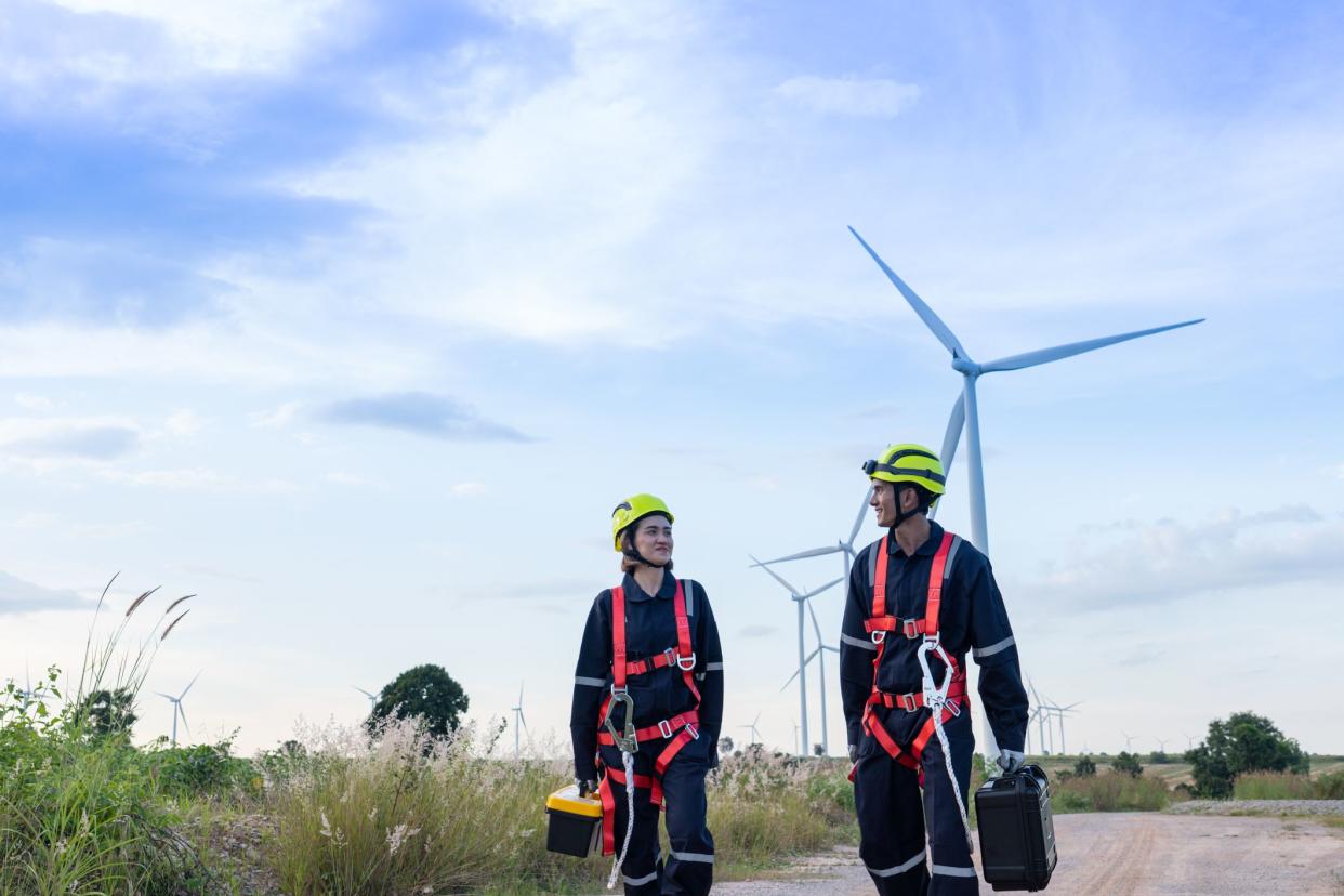 Male and female engineers in uniform, red safety bells, yellow hats, non-slip gloves for working at height walk into a wind turbine farm to perform maintenance. Clean energy from nature.