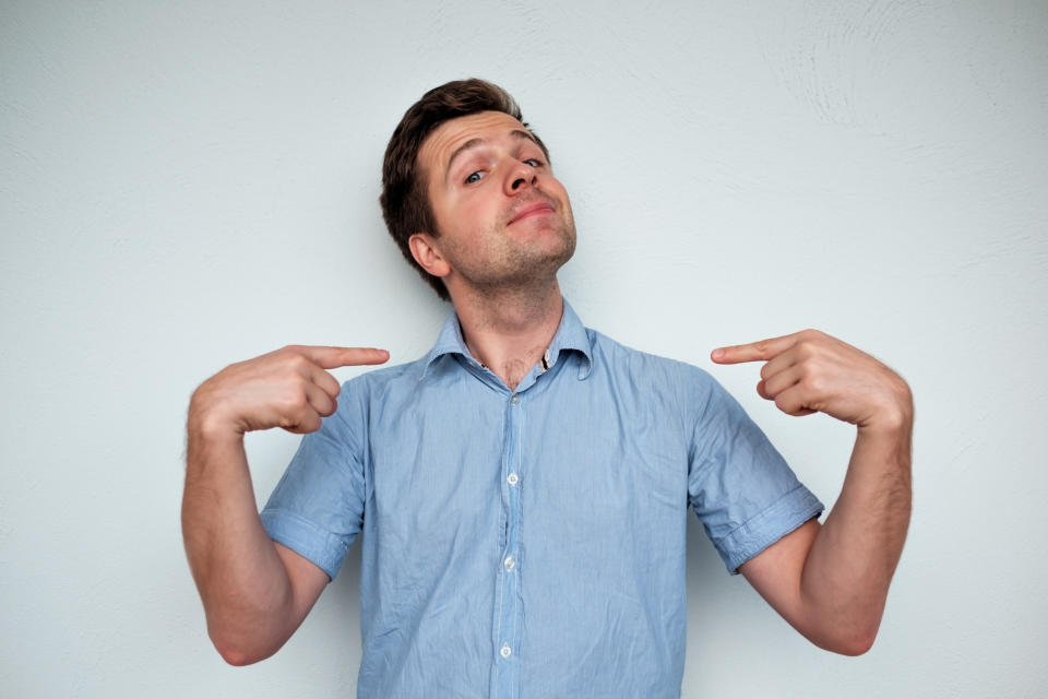 Man standing against gray background, pointing toward himself with arrogant expression