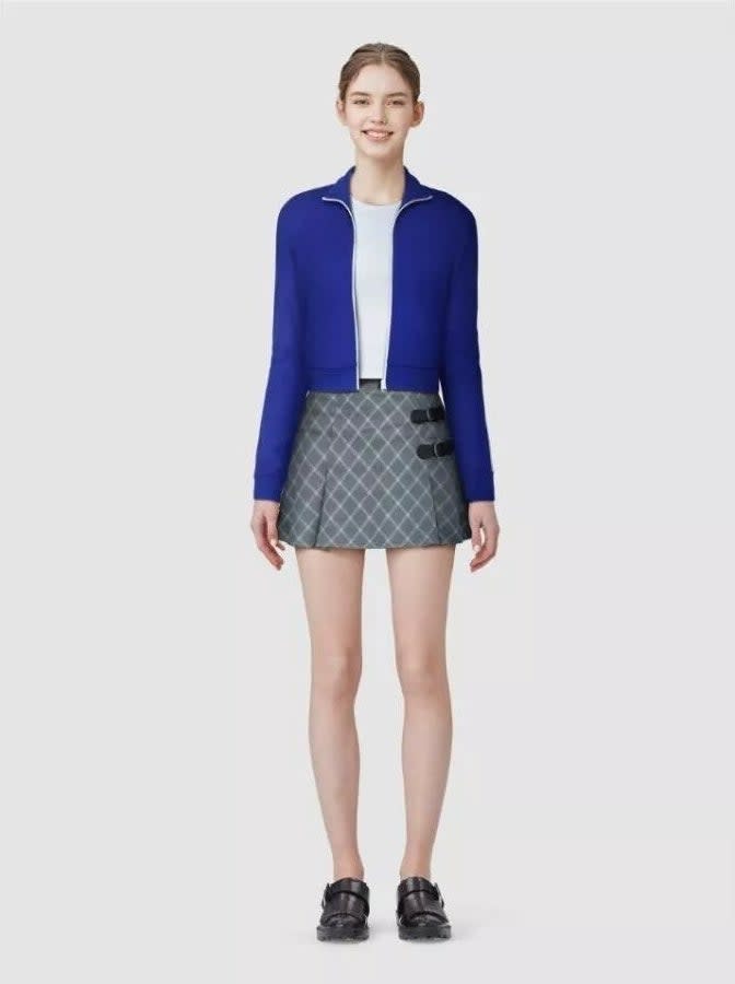 A model in a blue jacket and patterned skirt with black shoes stands smiling