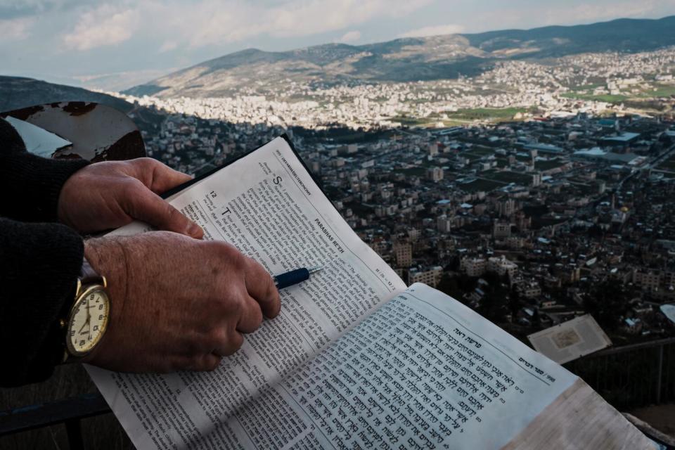 A hand pointing to a passage in English in the Torah, and a sprawling community in the valley below