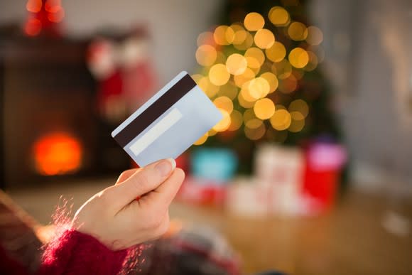 A person holds a credit card in the air in front of a Christmas tree surrounded by presents.