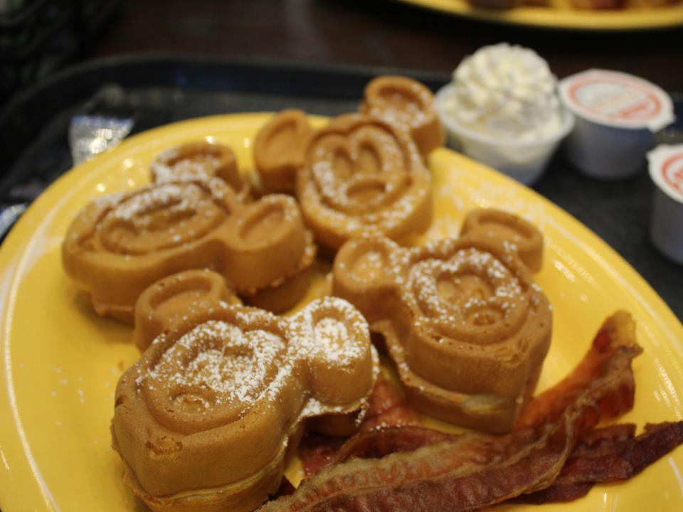 Mickey-shaped waffles on a yellow plate with bacon taken at a Disney park breakfast.