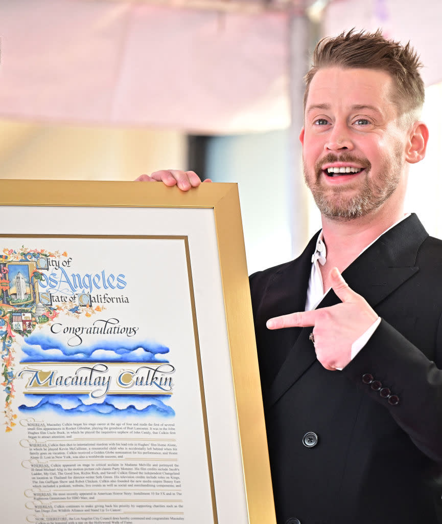 Man in suit holding a framed certificate with the name "Macaulay Culkin" on it, smiling and pointing at the text