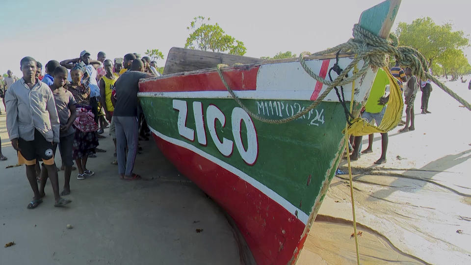 In this video grab provided by Television Mozambique, people stand on Monday, April 8, 2024, next to a boat that local media say capsized on Sunday off Mozambique's northern coast in Nampula province. The makeshift ferry reportedly was overcrowded with residents fleeing a feared cholera outbreak when it capsized, killing at least 98 people including children, media reports say. (Television Mozambique via AP)