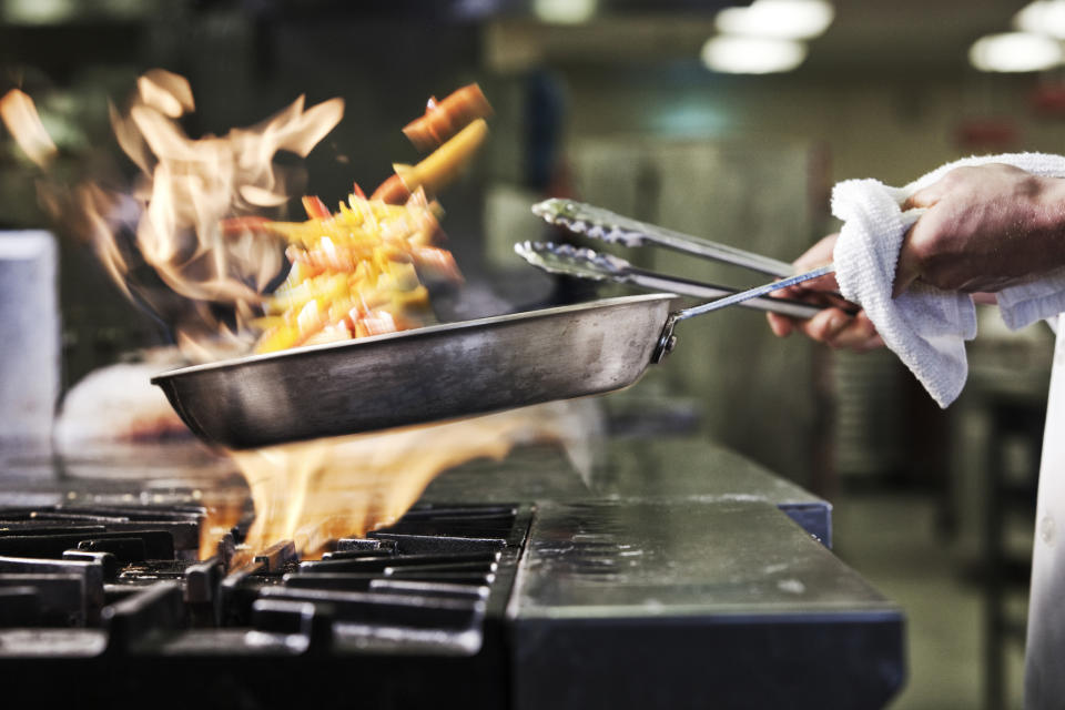 Close-up of chef’s hands holding a saute pan to cook food, flambeing contents. Flames rising from the pan. (Image: Getty)