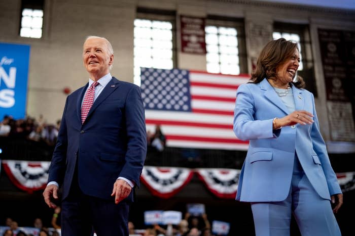 Joe Biden in a suit with a red tie and Kamala Harris in a light blue pantsuit, standing in front of a crowd with an American flag in the background