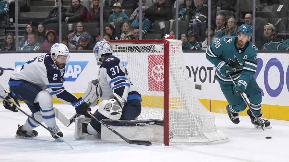 San Jose Sharks defenseman Mario Ferraro (38) skates with the puck behind the net, as Winnipeg Jets defenseman Dylan DeMelo (2) and goaltender Connor Hellebuyck (37) watch during the first period of an NHL hockey game in San Jose, Calif., Thursday, Jan. 4, 2024. (AP Photo/Jeff Chiu)