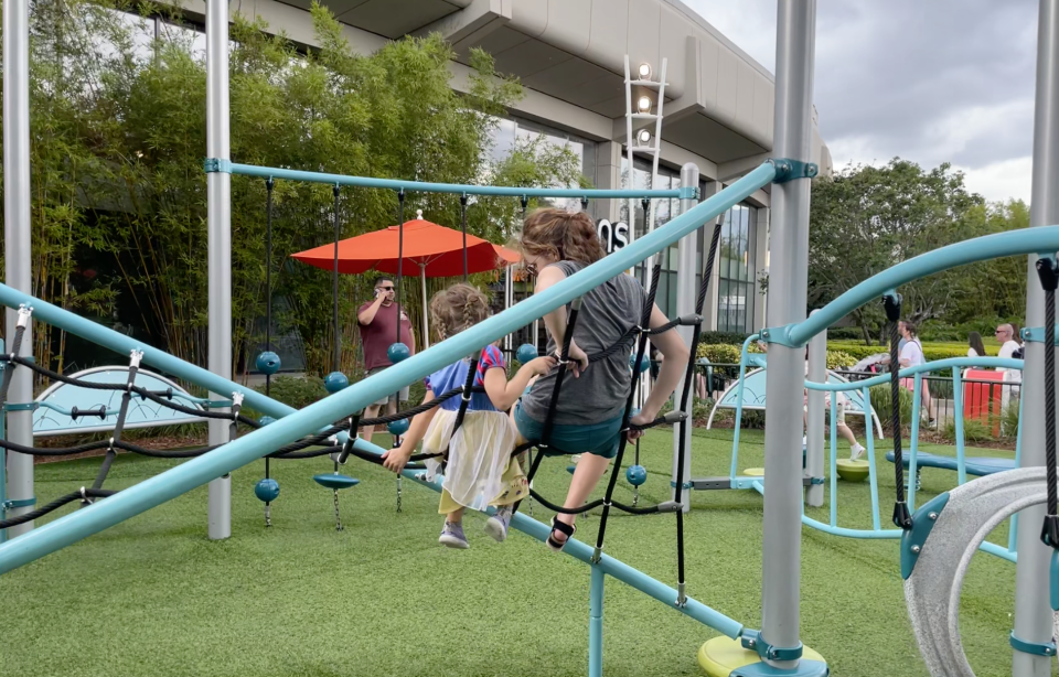 Two children swing together on a play structure outside, while adults in the background watch and take photos
