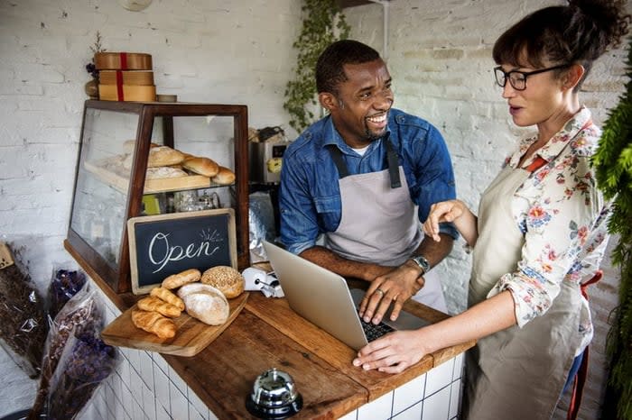 A cafe owner standing behind the counter with a laptop and joking with his employee.