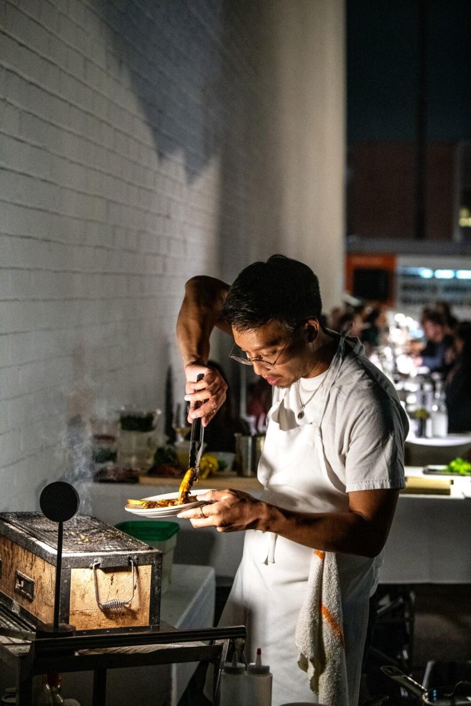 A man in white plates food cooked on a portable cooktop in an alley.