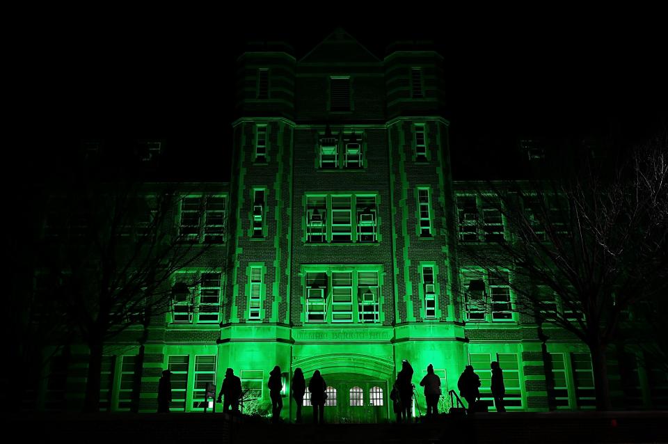 People stop and visit outside Michigan State University’s Berkey Hall, which was illuminated green as a tribute to those affected by the mass shooting a year ago, on Tuesday, Feb. 13, 2024, in East Lansing.