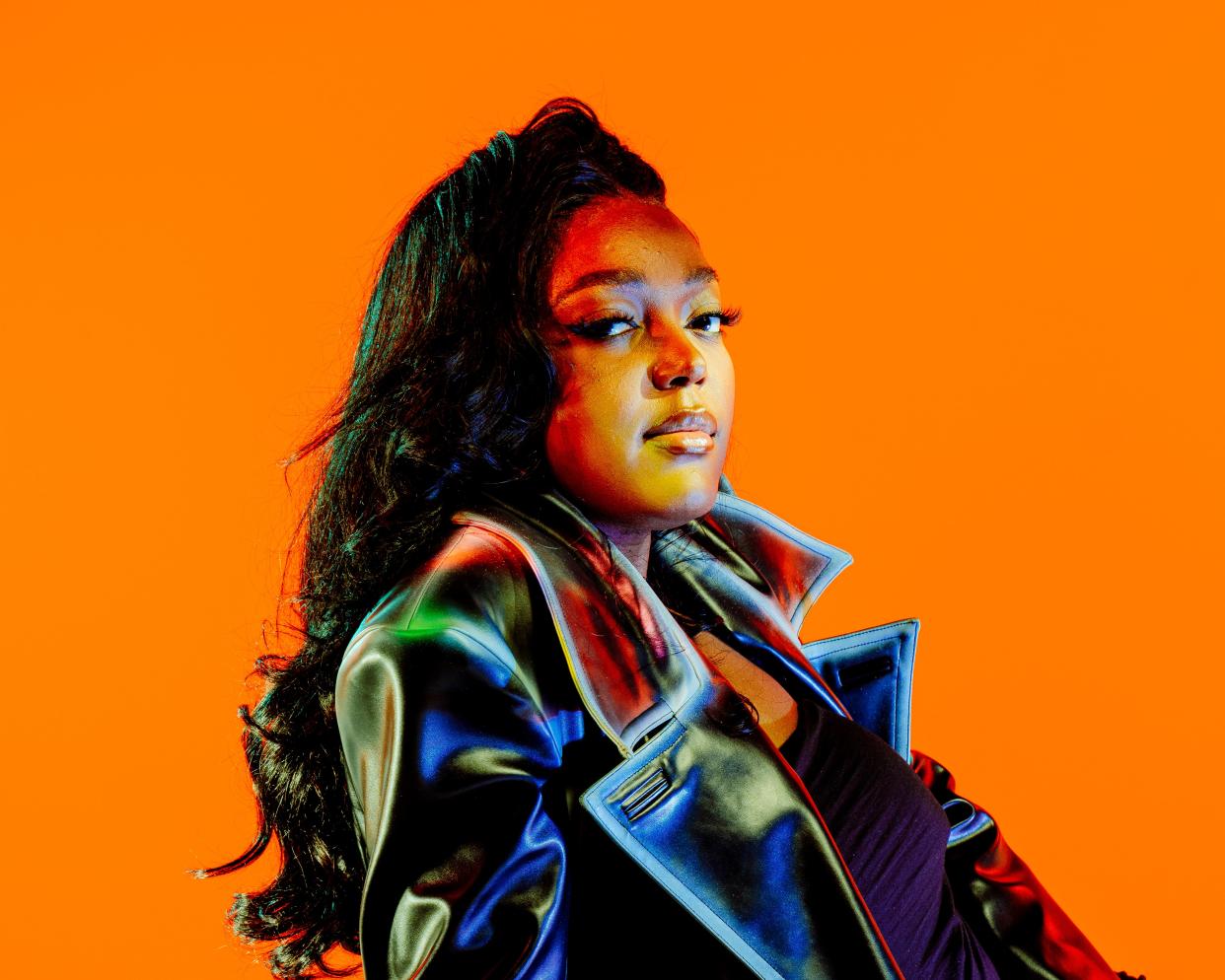 Anifa Mvuemba poses in a black leather coat against a neon orange backdrop.