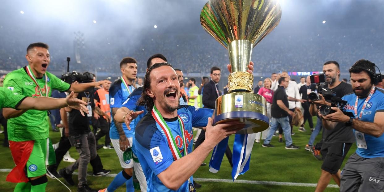 soccer player in blue uniform holding massive trophy on pitch