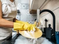 A person wearing a gingham apron and yellow rubber gloves is standing at the sink washing dishes.