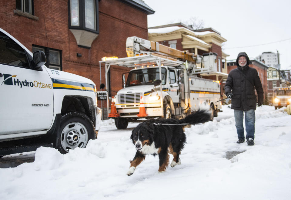 A man walks his dog past Hydro Ottawa utility vehicles responding to power outages in parts of Ottawa's Golden Triangle neighborhood, as a winter storm warning is in effect, on Friday, Dec. 23, 2022. (Justin Tang /The Canadian Press via AP)