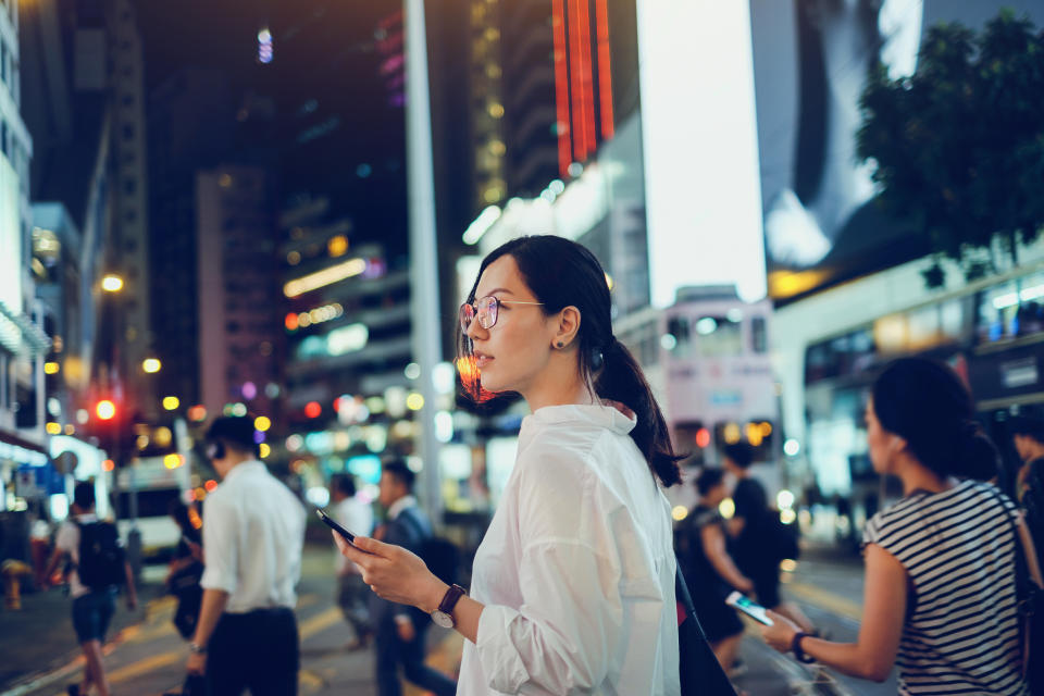Beautiful Asian woman using mobile phone while crossing road in busy downtown city street at night (Photo: d3sign via Getty Images)