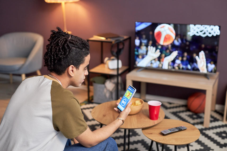 A person with braided hair looks at a smartphone while watching a basketball game on TV at home. Nearby are a red cup, remote, and snacks on a table