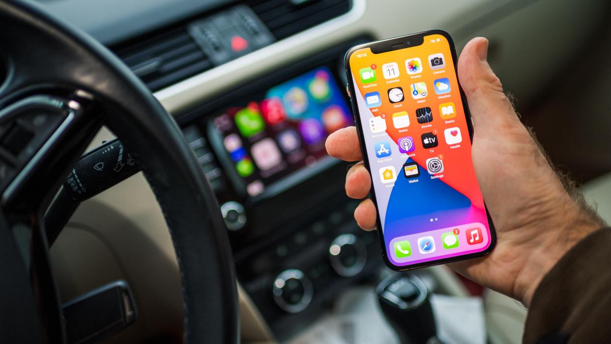  Man's hand holding iPhone in car with CarPlay screen behind. 