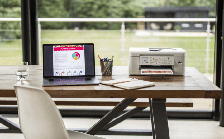 The Brother MFC-J1010DW Wireless All-in-One Inkjet Printer on a desk next to a laptop.