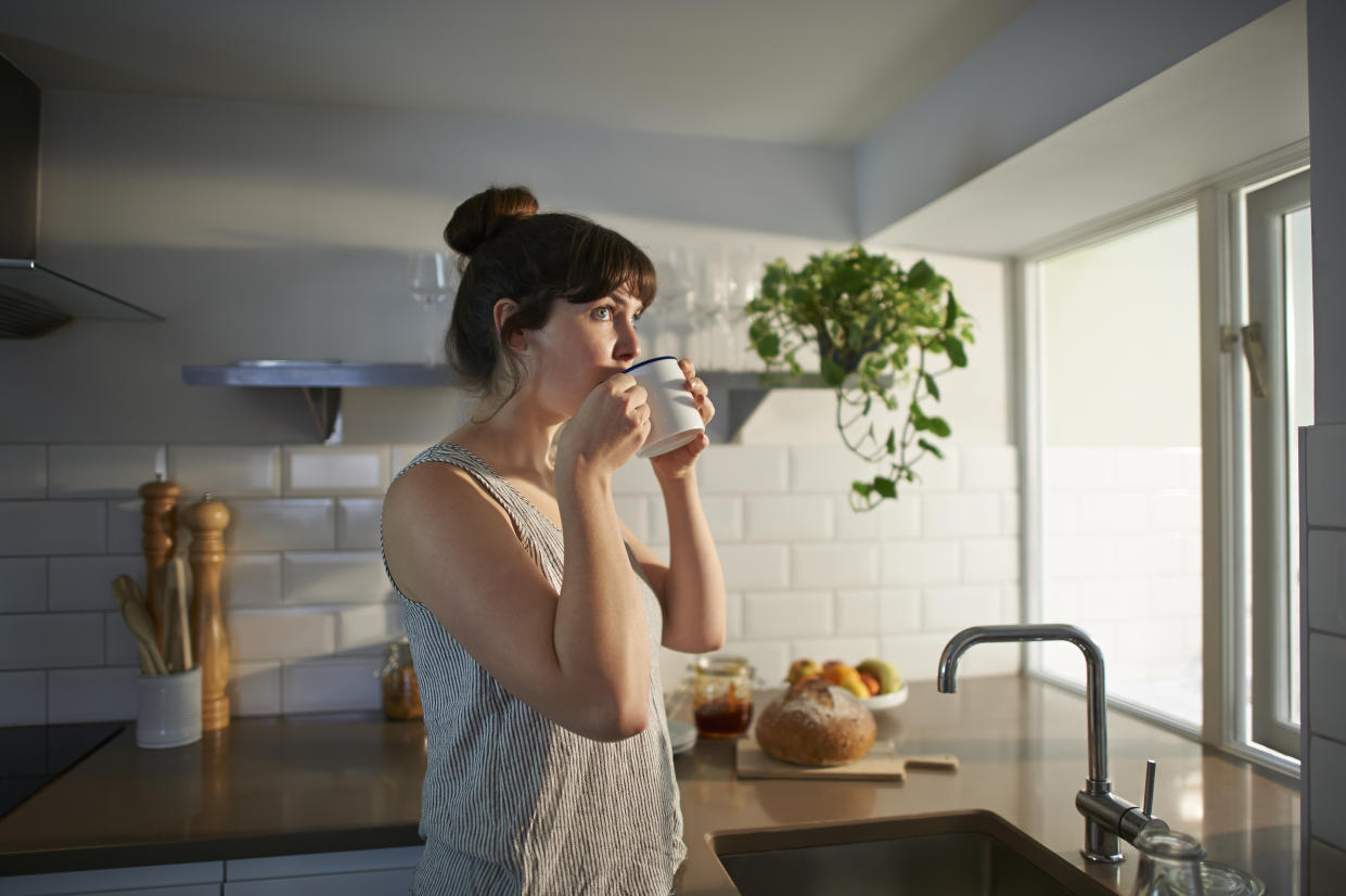 A woman standing in her kitchen drinks from a mug. single