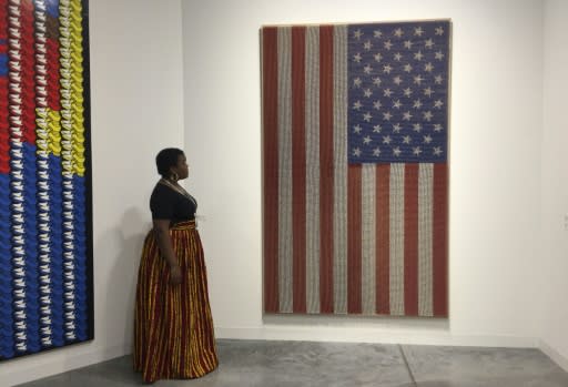 An exhibitor stands next to a piece by Rob Pruitt titled "Pledge Allegiance (Flammable)," which is made of sticks hand-painted to look like matches