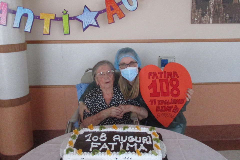 In this photo taken on Wednesday, June 3, 2020, Fatima Negrini, left, is flanked by nurse Laura Catena holding a cut out heart reading in Italian " Fatima 108, we love you " while posing in front of a cake celebrating her 108th birthday, at the Anni Azzurri San Faustino nursing home, in Milan, Italy. Fatima Negrini has joined the ranks of the very few centenarians who have recovered from COVID-19. (RSA Anni Azzurri San Faustino via AP)