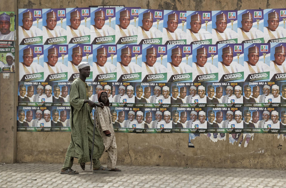 A child leads a blind man as they walk past a wall of campaign posters in Kano, in northern Nigeria, Saturday, Feb. 16, 2019. A civic group monitoring Nigeria's now-delayed election says the last-minute decision to postpone the vote a week until Feb. 23 "has created needless tension and confusion in the country." (AP Photo/Ben Curtis)