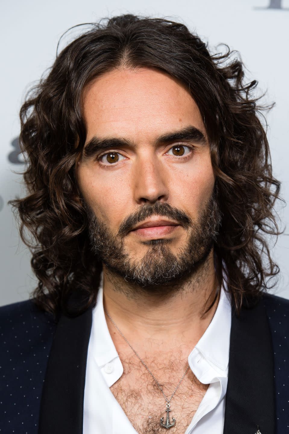 russell brand pictured in 2017, he has long hair and a beard and looks into the camera