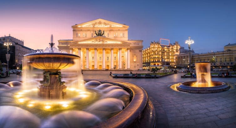 A stately theatre lit at night with fountains outside.