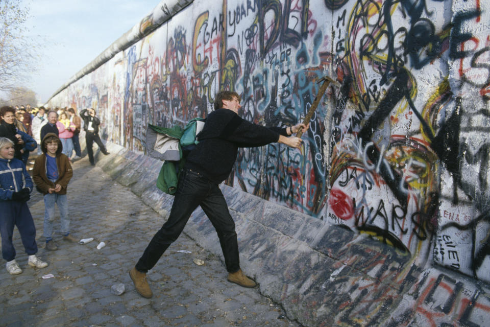 A man strikes the Berlin Wall with a pickax as people a few feet away watch.