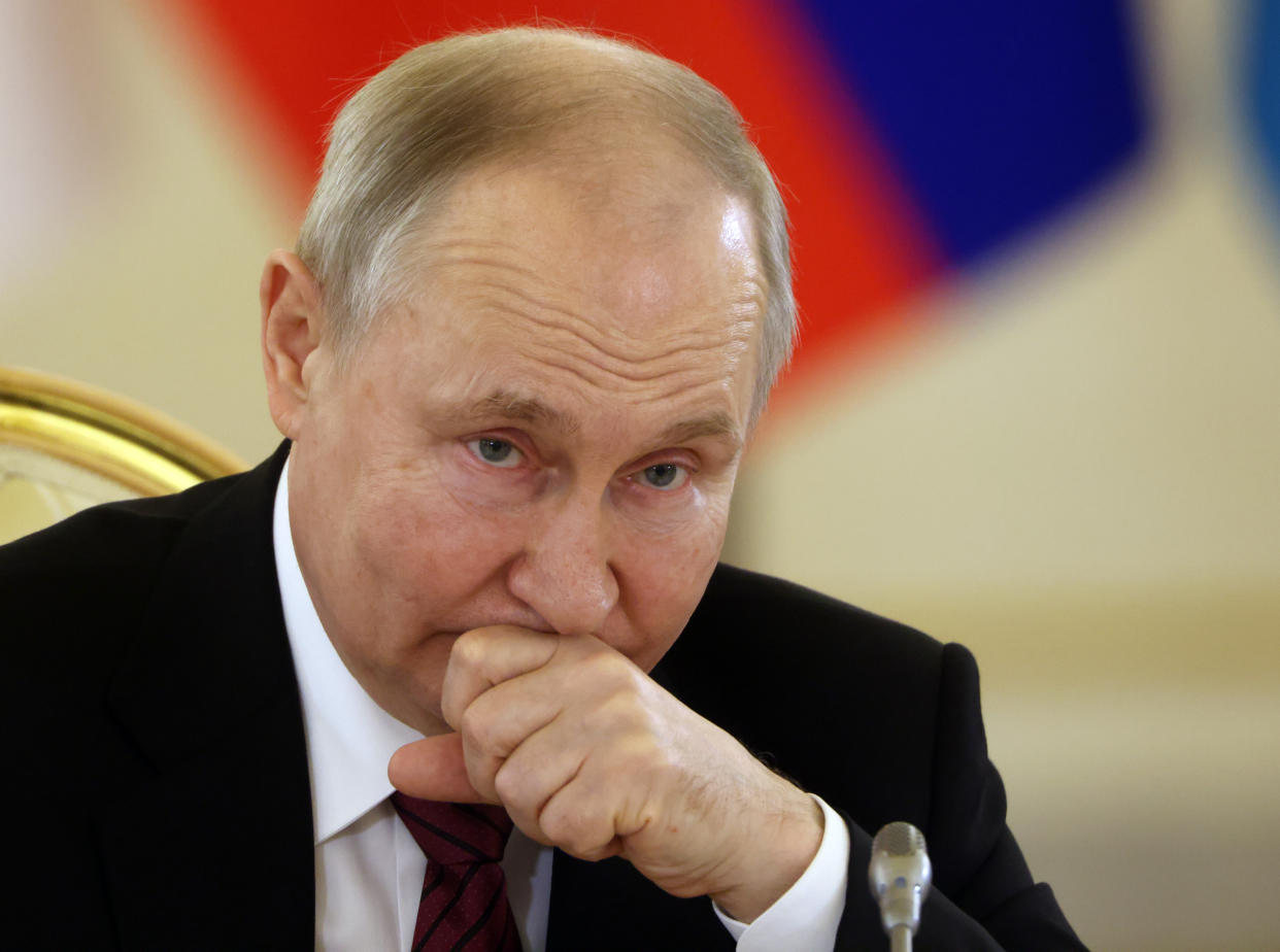 Russian President Vladimir Putin, sitting in a gilt chair, covers his mouth with his hand.