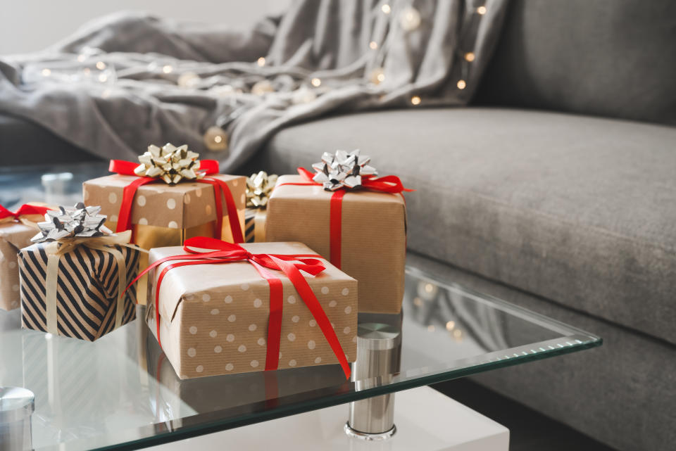 Gift boxes wrapped in craft paper on glass coffee table in the living room, sofa bed with blanket and decorative lights as background. Christmas presents