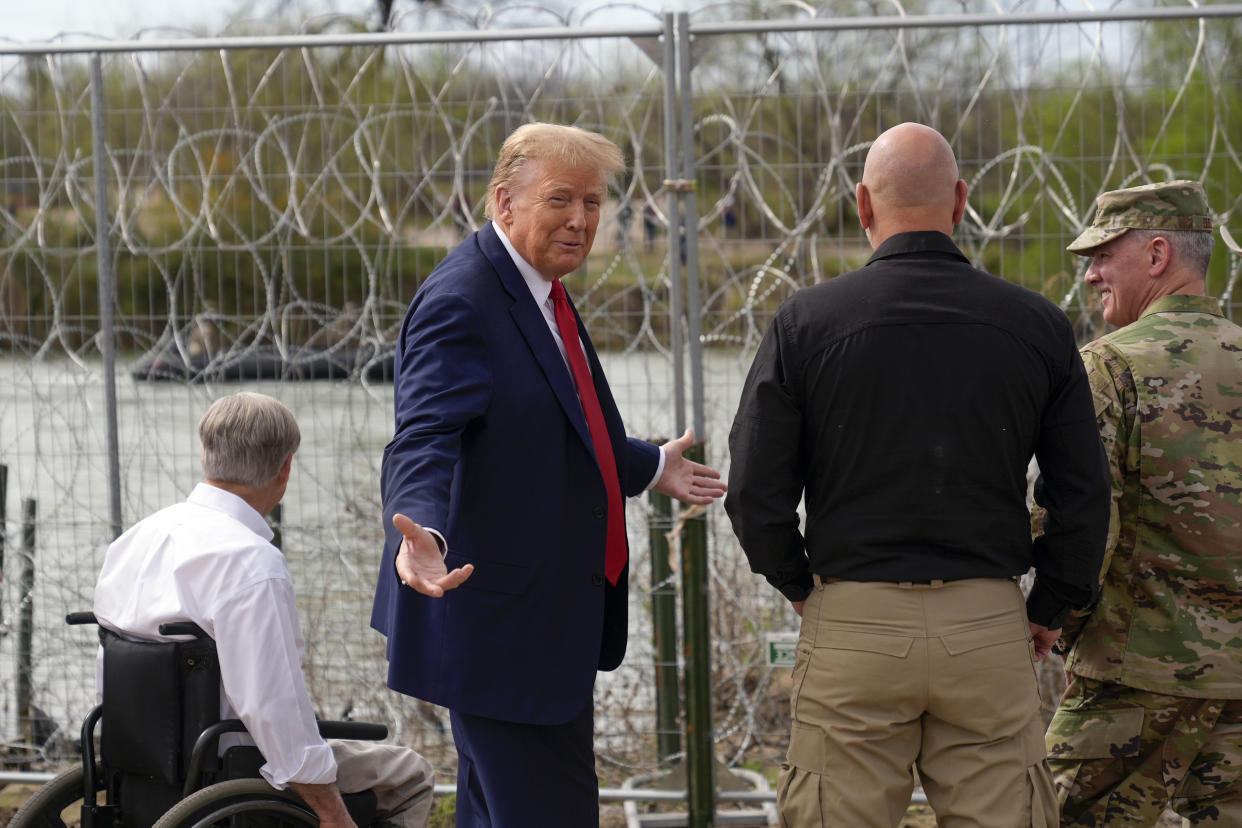 Donald Trump stands next to a few people by a barbwire fence near a riverbank.