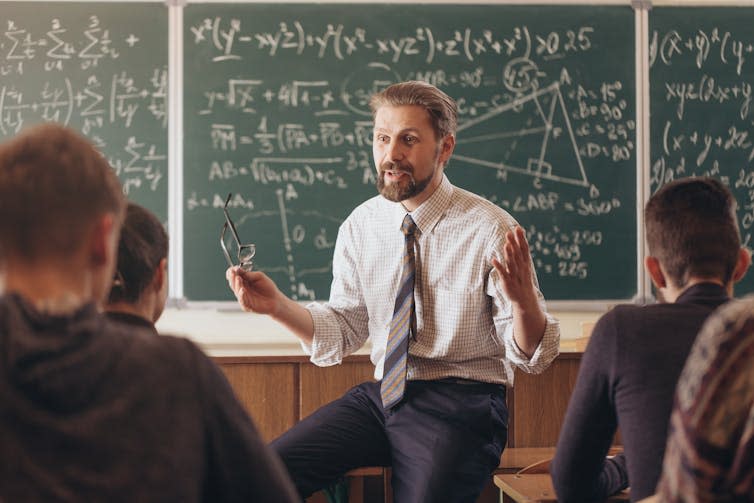 A male teacher sitting at the front of the classroom addressing students, with a chalkboard full of equations behind him