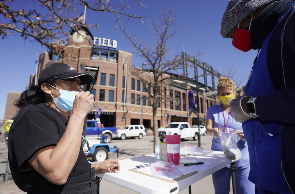 Martha Bravo, left, self-administers a swab COVID-19 test under the guidance of Josh Copeland at a table set up outside the main gate of Coors Field as fans return for the first time to the ballpark for a baseball game between the Los Angeles Dodgers and the Colorado Rockies Thursday, April 1, 2021, in Denver. Community Wellness of America was offering the service to fans as they entered the ballpark. (AP Photo/David Zalubowski)