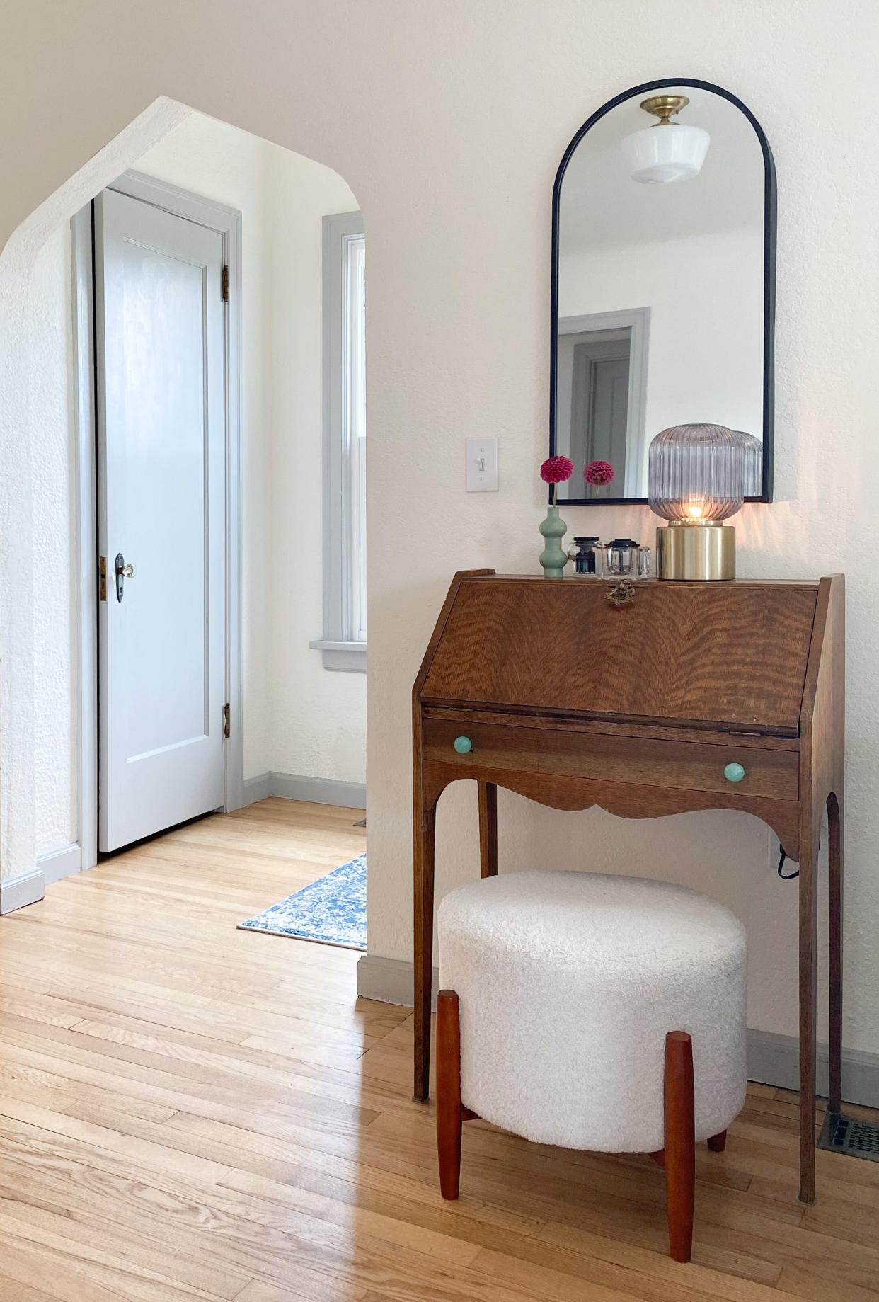 A modern mirror, bud vase, lamp and ottoman update the vintage desk near the entryway.