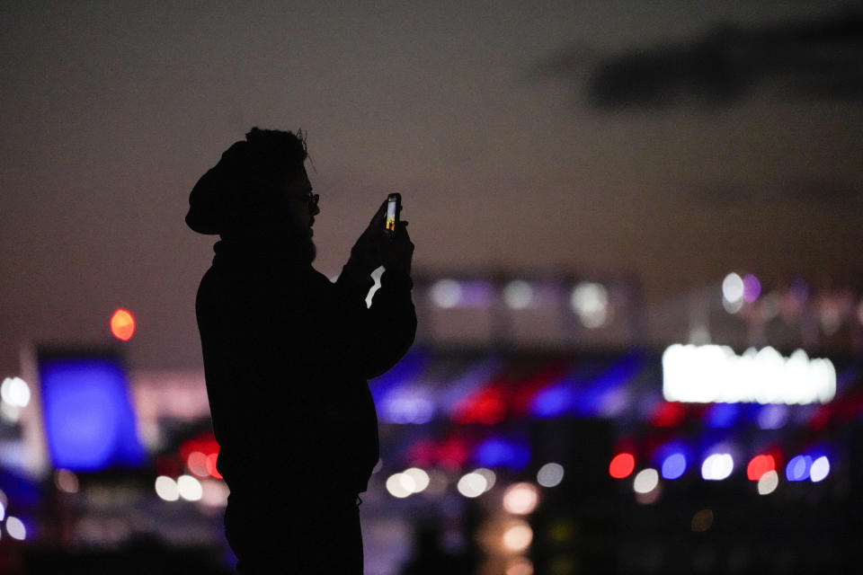 A man uses a cell phone to capture images at Federal Hill Park with M&T Bank, the home stadium of the Baltimore Ravens, giving a backdrop in the colors of the Buffalo Bills in support of safety Damar Hamlin, Wednesday, Jan. 4, 2023, in Baltimore. Hamlin was taken to the hospital after collapsing on the field during an NFL football game against the Cincinnati Bengals on Monday night. (AP Photo/Julio Cortez)