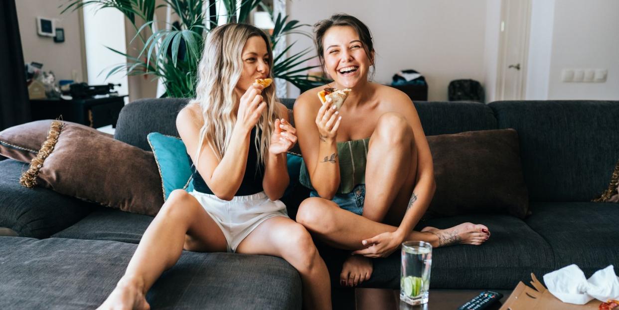 two lesbian women watching tv at home in living room and eating pizza