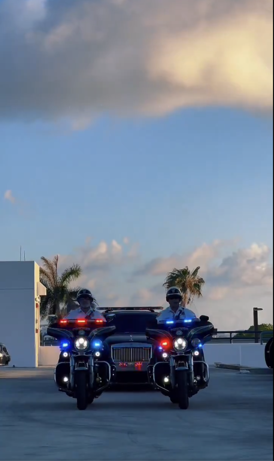 Two police officers ride motorcycles with flashing lights, leading a black car on a rooftop against a backdrop of palm trees and clouds