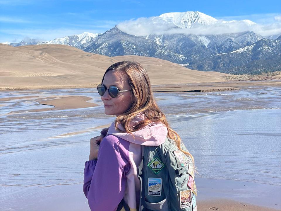 Emily, wearing a multi-colored jacket and a backpack with patches, looks over her shoulder in Great Sand Dune National Park. Behind her are snow-capped mountains.