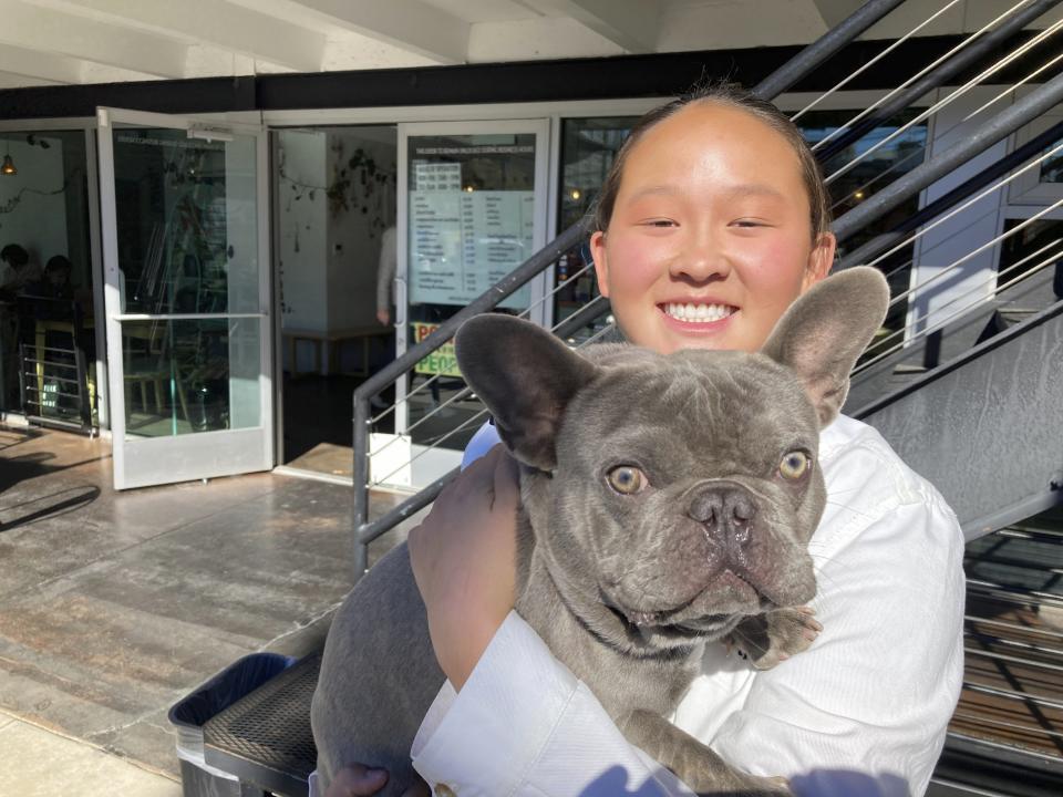 U.S. moguls skier Kai Owens poses with her French bulldog, Mochi, outside a coffee shop in Denver in this photo taken on Oct. 29, 2021. As an infant, she was abandoned at a town square in a province of China. Taken to an orphanage, she was adopted by a couple from Colorado at 16 months. Now 17, Owens is on the verge of earning a spot in moguls for the Winter Games in Beijing. It's a return to China she's long thought about. (AP Photo/Pat Graham)