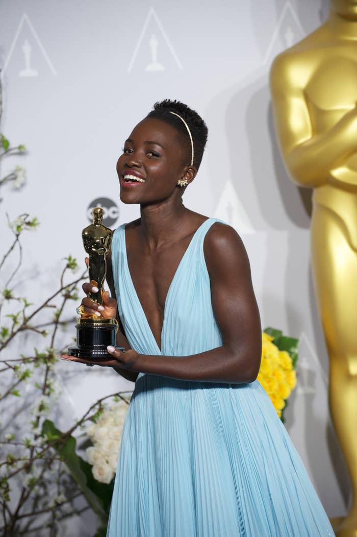 Lupita Nyong'o poses in a pale blue dress while smiling and holding an Oscar