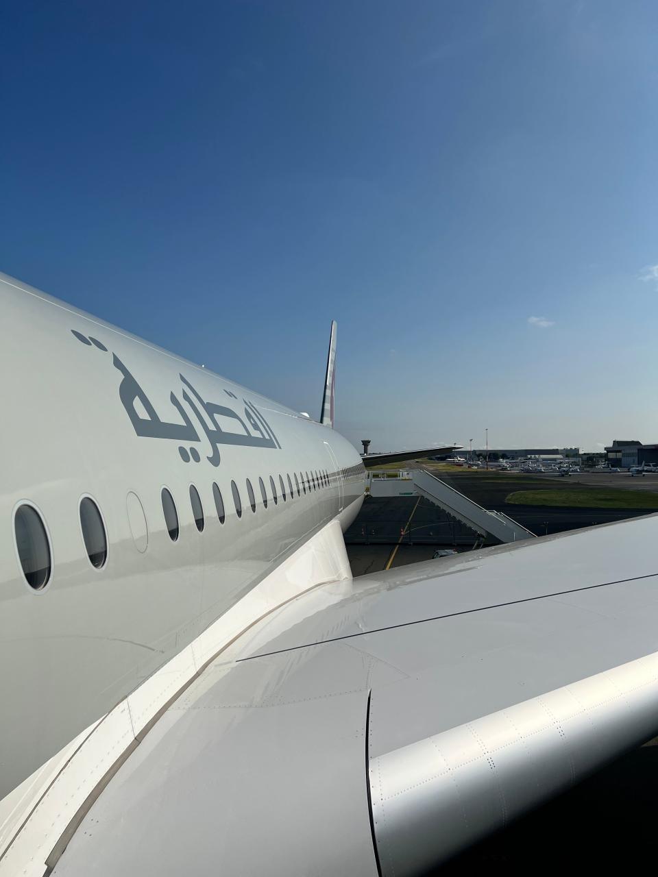 The side of a Qatar Airways A350 with Arabic writing on it, viewed from the center door, part of the wing and a rear staircase are visible