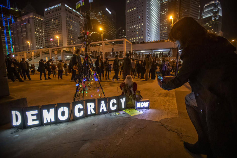 A protester takes a photo of a Christmas tree and Santa Claus figure next to a sign reading "Democracy Now" at a rally in Hong Kong, Thursday, Dec. 12, 2019. Protesters in Hong Kong have written hundreds of Christmas cards for detainees jailed in the city's pro-democracy movement. At a rally on Thursday night, protesters promised on the cards that detainees won't be forgotten as they face spending the festive season behind bars. (AP Photo/Mark Schiefelbein)