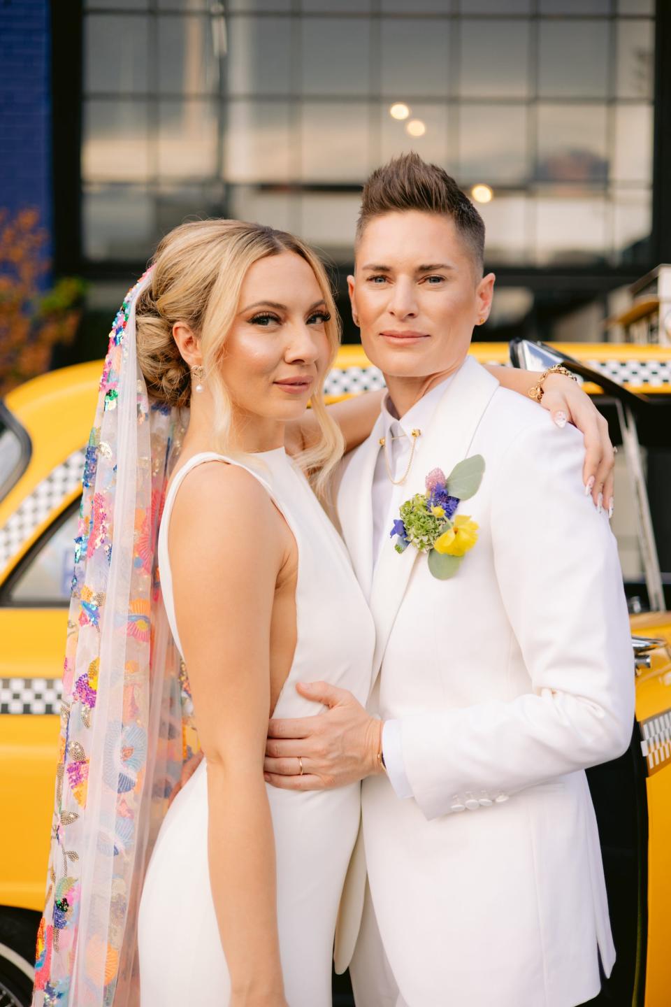 A bride in a white dress and rainbow veil and a bride in a white suit embrace in front of a taxi.