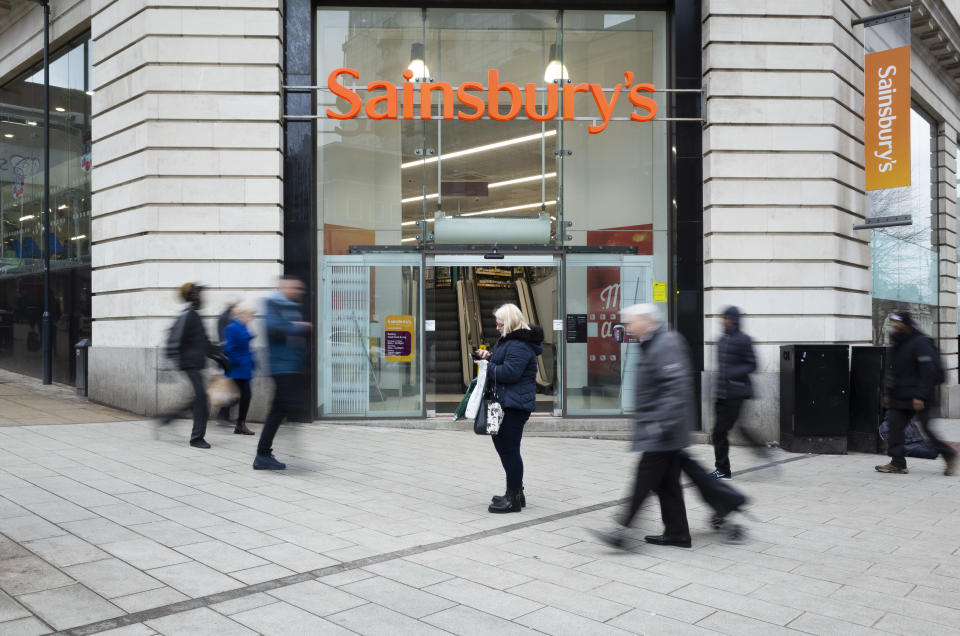 A lone woman looks at her phone while other members of the public, some of them with carrier bags and wearing winter clothes, pass her by outside the doors of a Sainsbury's, one of Britain's largest supermarkets, amid declining consumer spending and a cost of living crunch in the country on 28th February, 2022 in Leeds, United Kingdom. The latest economic analysis points towards huge blows in consumer spending and confidence in the UK as energy bills and the cost of basic goods rise in tandem, with inflation predicted to peak at nearly 8% and float above 6% for most of the year. (photo by Daniel Harvey Gonzalez/In Pictures via Getty Images)