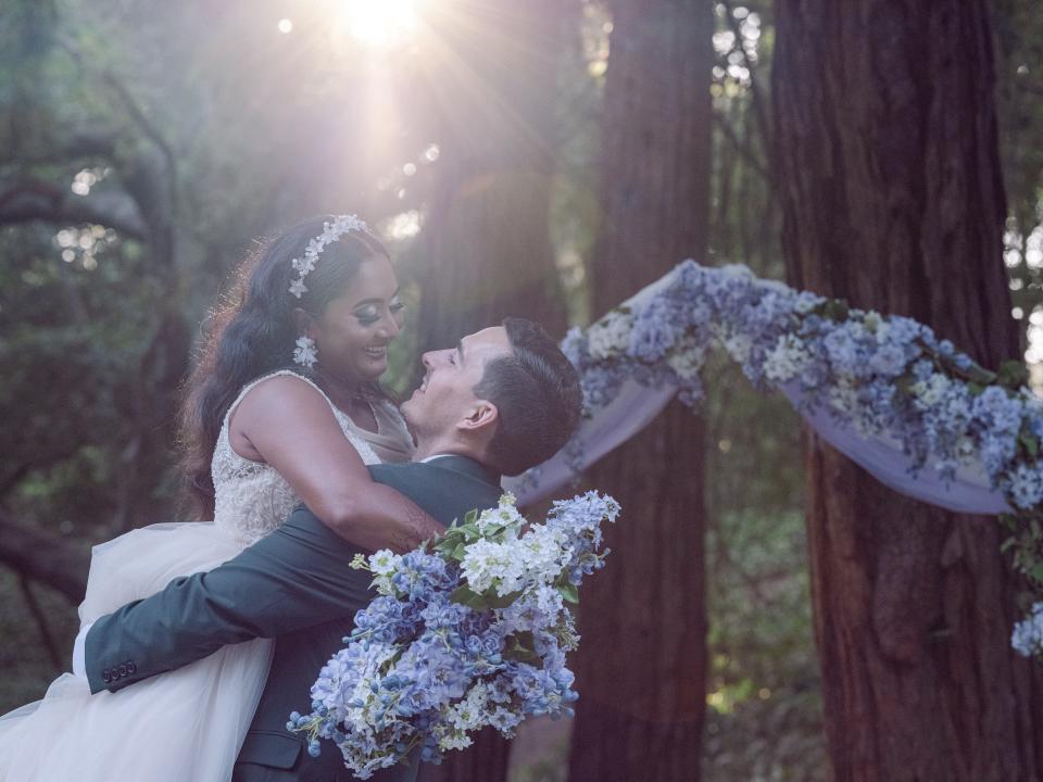 The groom is lifting the bride up and they are smiling at each other in front of the purple floral backdrop they used for their outdoor wedding ceremony. The bride is holding a matching purple bouquet.